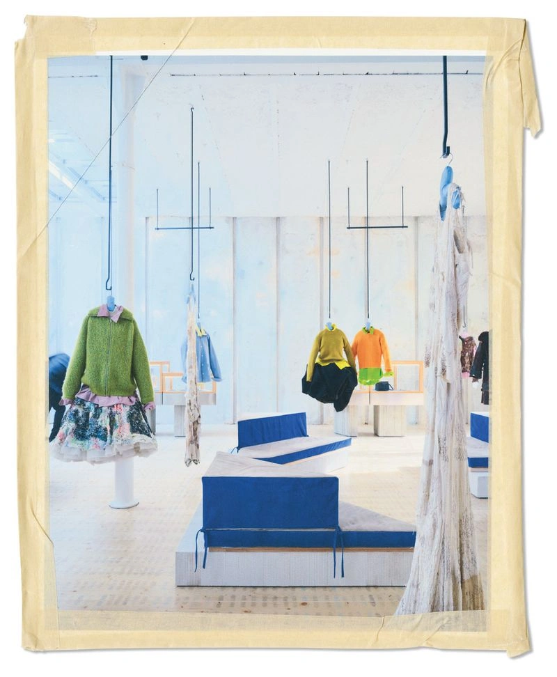 Interior of an American Vogue fashion editorial space featuring garments suspended from ceiling hooks and blue and white seating platforms.