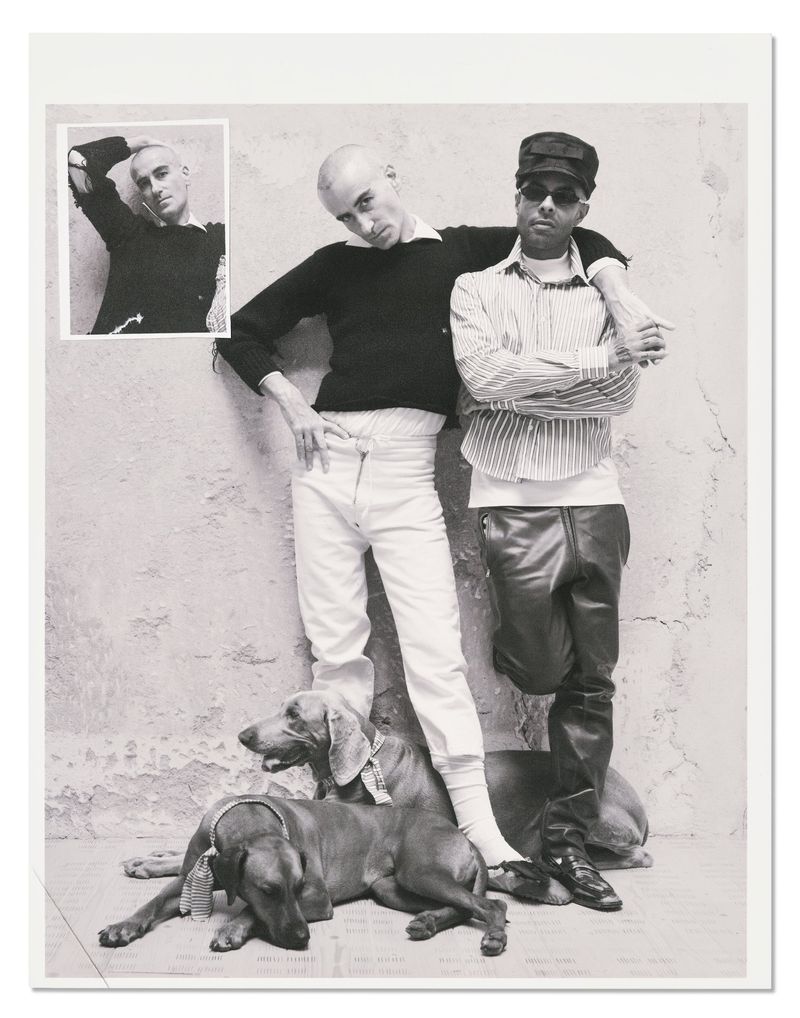 Black and white editorial photograph of two men posing with two dogs against a textured wall with an inset image