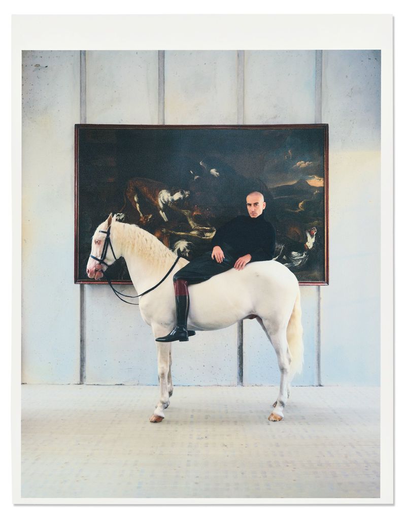 Bald man seated atop a white horse indoors against a wall with a large dark painting, photographed by Pablo Di Prima.