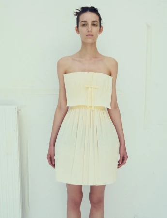 A model wearing a strapless pale yellow mini dress standing against a plain light wall for Dazed Magazine.