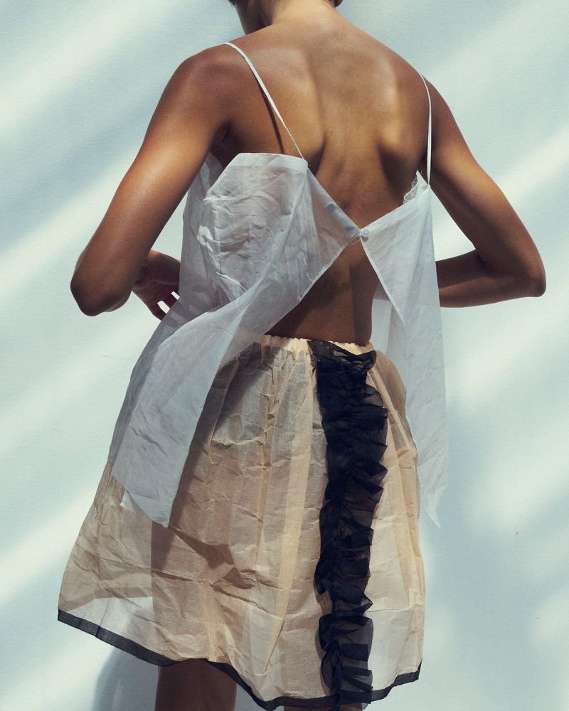 Model seen from behind wearing a white sheer top and a cream skirt with a black ruffle detail against a plain white wall.