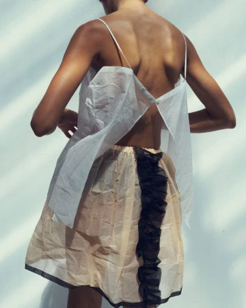 Model seen from behind wearing a white sheer top and a cream skirt with a black ruffle detail against a plain white wall.