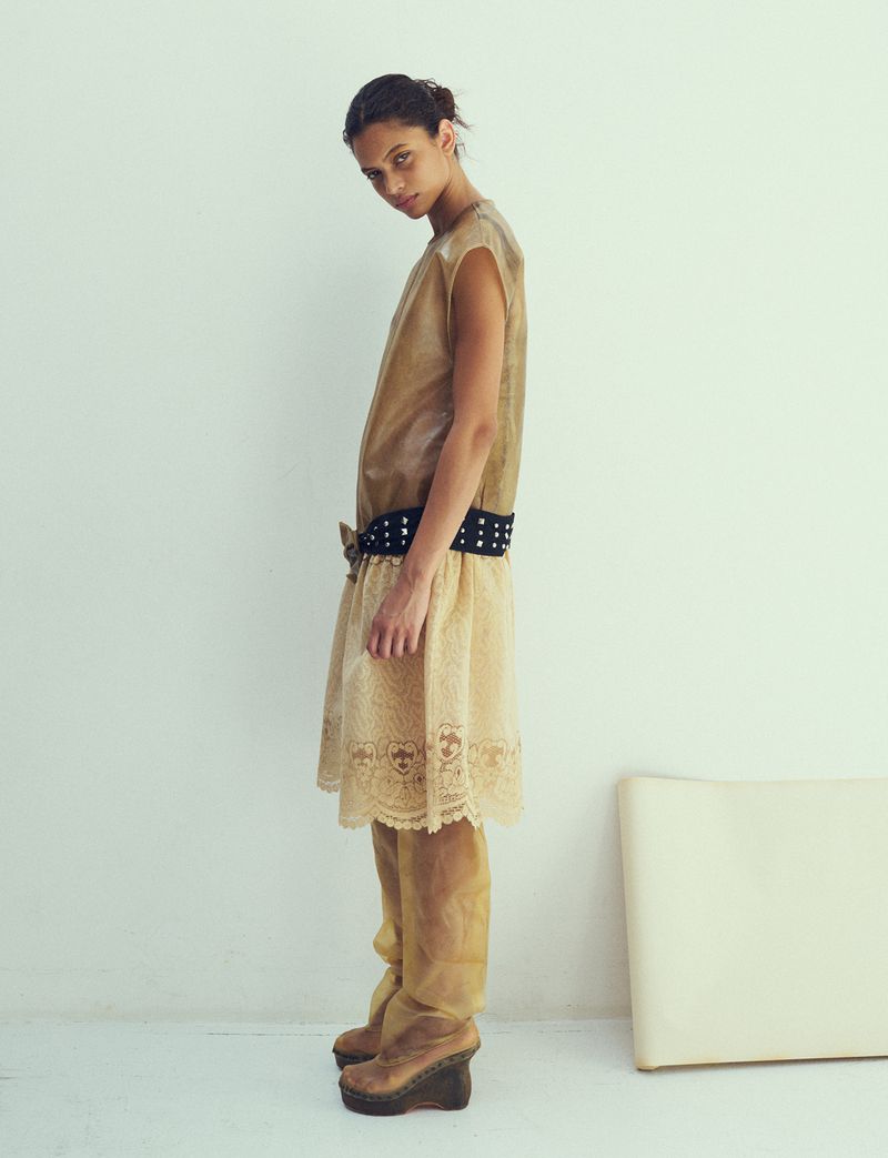 Model in beige layered dress and studded belt standing against a plain white background for Dazed Magazine editorial