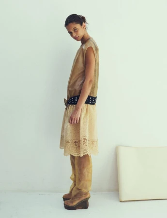 Model in beige layered dress and studded belt standing against a plain white background for Dazed Magazine editorial