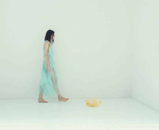 Model in a sheer pale blue dress walking barefoot across a white studio floor past a yellow plastic bag.