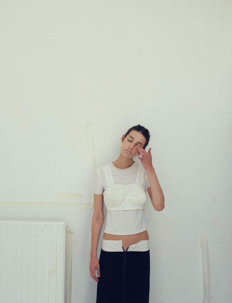 Young woman wearing a white top with a bustier overlay and dark skirt standing against a plain white wall.