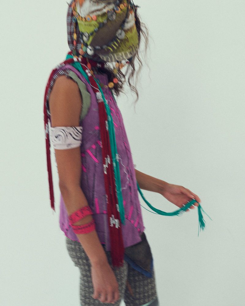 Model in layered purple sleeveless top and patterned leggings wearing a decorated headscarf against a white background.
