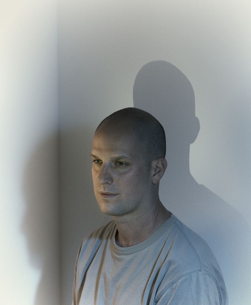 Portrait of a bald man with a serious expression casting a prominent shadow on a plain wall, photographed by Nadav Kander for WSJ.