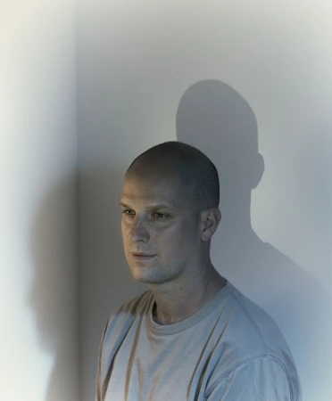Portrait of a bald man with a serious expression casting a prominent shadow on a plain wall, photographed by Nadav Kander for WSJ.