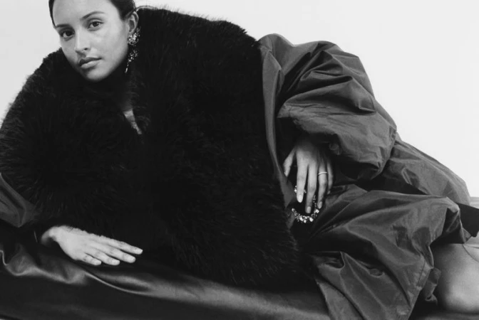 Woman in a dark faux fur coat and voluminous puffer jacket reclining against a white background