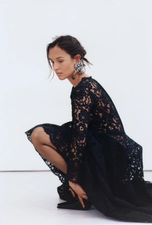 Woman in black lace dress crouching against a plain white background wearing an ornate earring