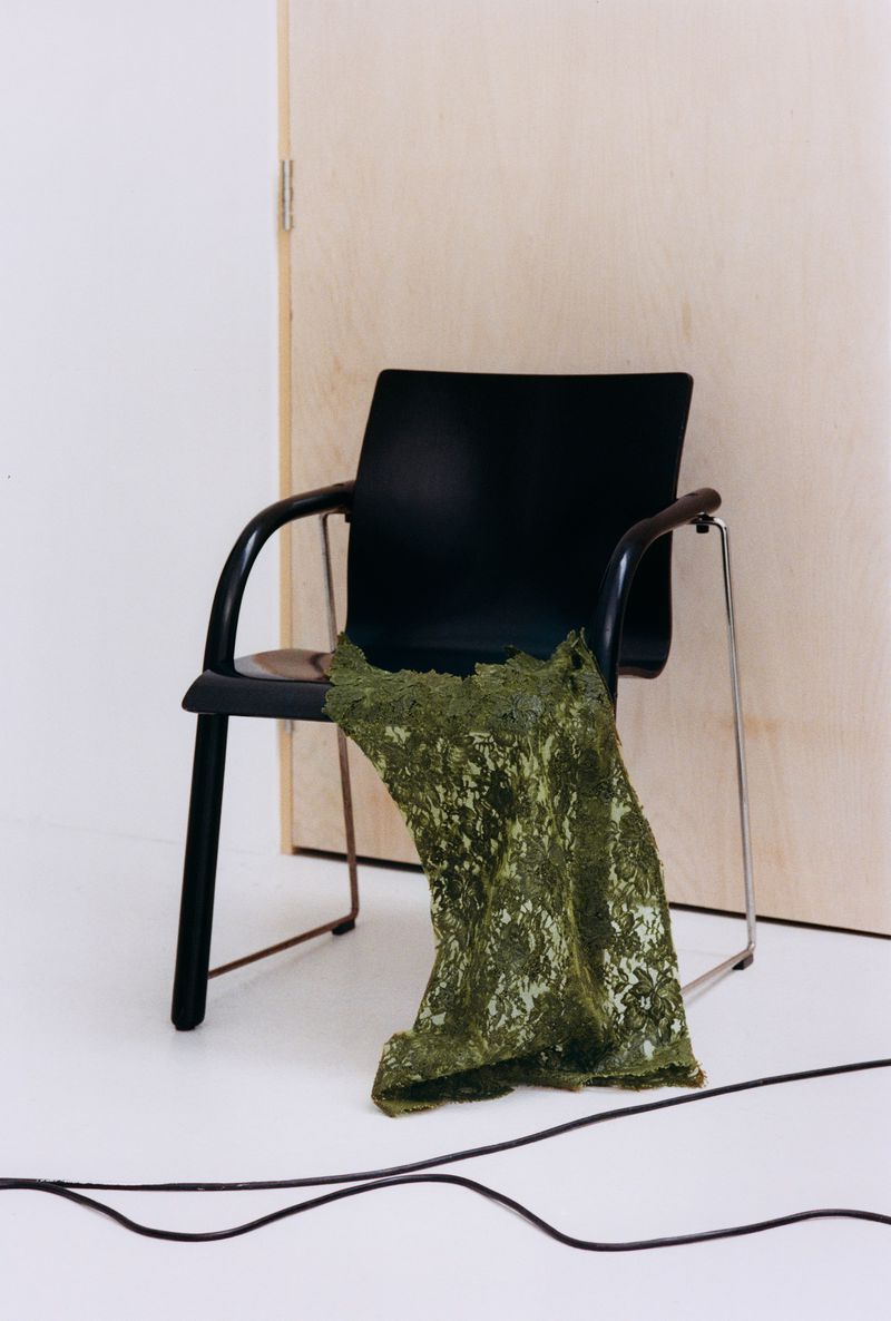 A black modern chair with a textured green lace fabric draped over it set against a wooden door and white floor