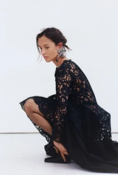 Woman in black lace dress crouching against a plain white background wearing an ornate earring