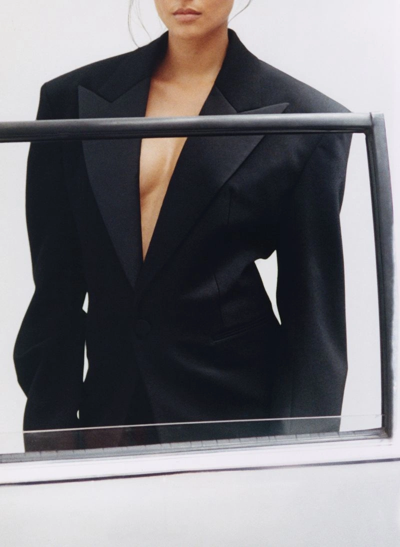 Model wearing a structured black blazer with satin lapels posed behind a dark framed glass barrier against a white background.