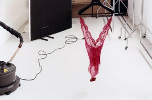 Red lace bodysuit hanging on a black hanger in a white studio with a steamer on the floor.