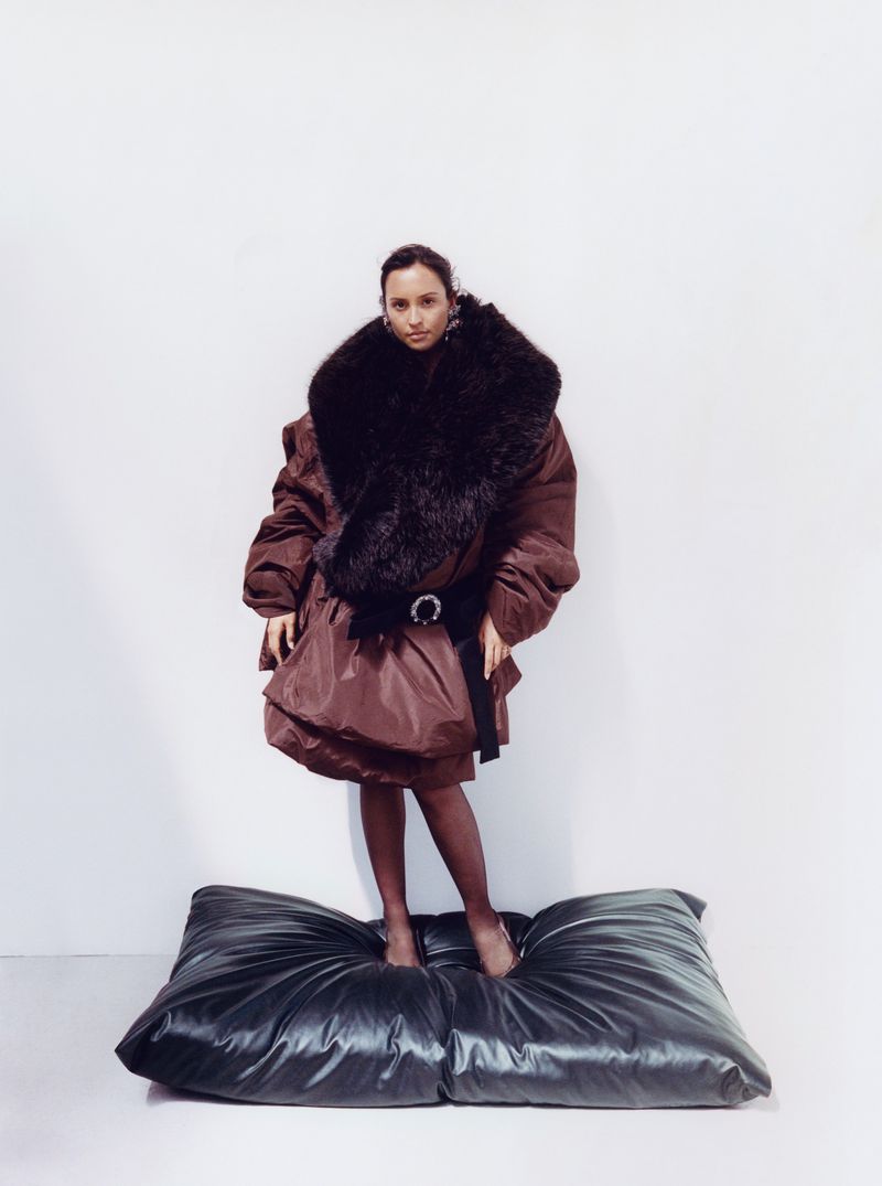 Model standing on a large dark cushion wearing a voluminous brown puffer coat with a massive faux fur collar against a plain white wall