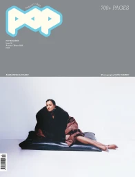 Woman in a black and brown puffer jacket reclining on a black leather cushion against a white studio background.