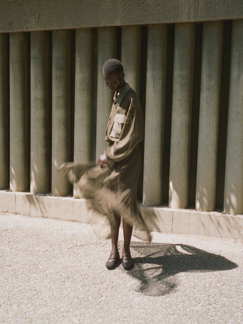 A model standing in front of concrete pillars wearing a utility shirt and sheer skirt, captured with motion blur.