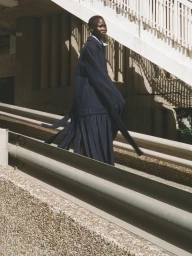 Model standing outdoors wearing a dark blue belted coat on an incline by a concrete structure