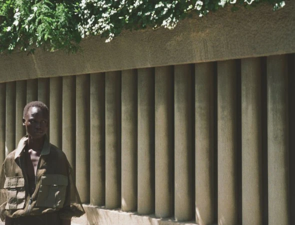 Model standing outdoors wearing a mesh utility jacket against a concrete colonnade with hanging greenery for Modern Road Travessera