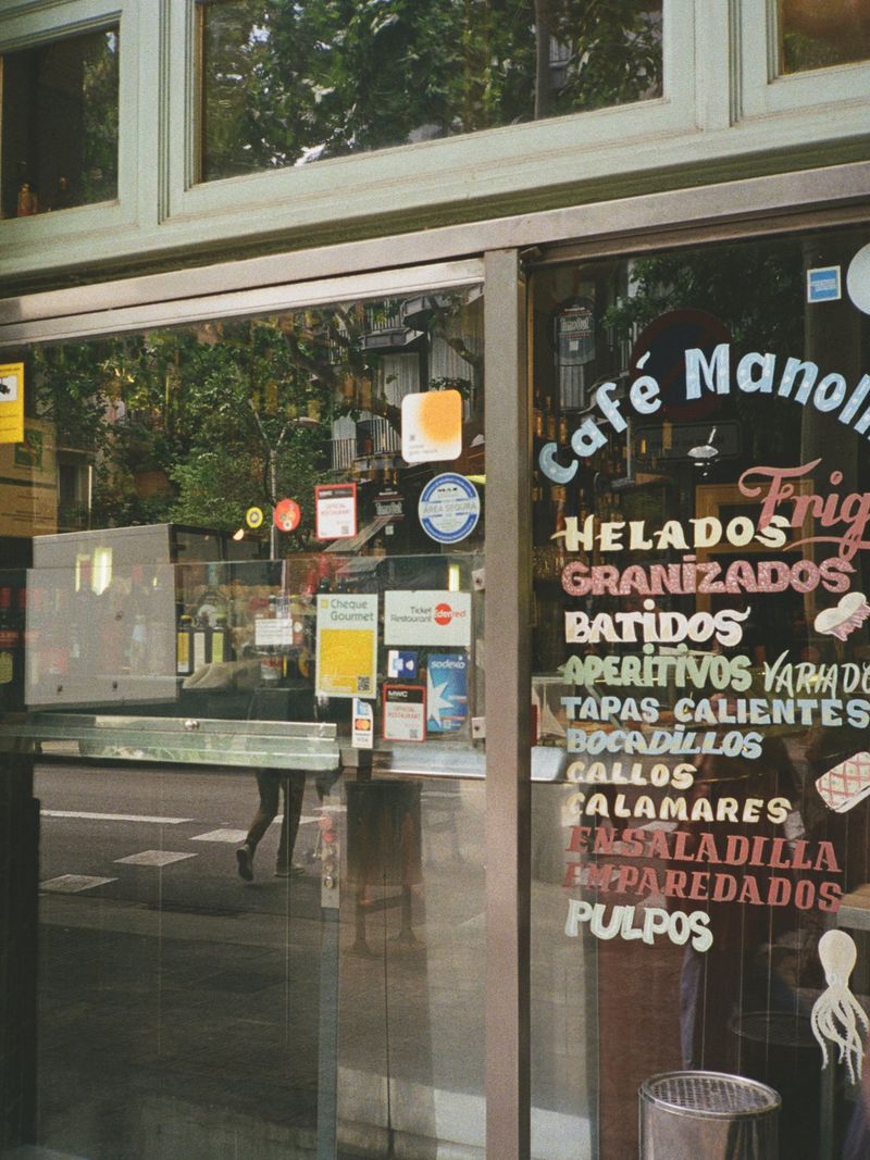 Storefront glass window of Cafe Manolo displaying painted menus and payment decals with street reflection