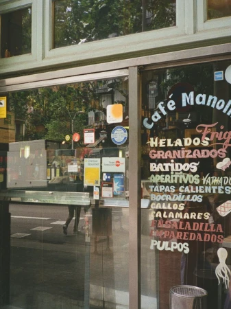 Storefront glass window of Cafe Manolo displaying painted menus and payment decals with street reflection