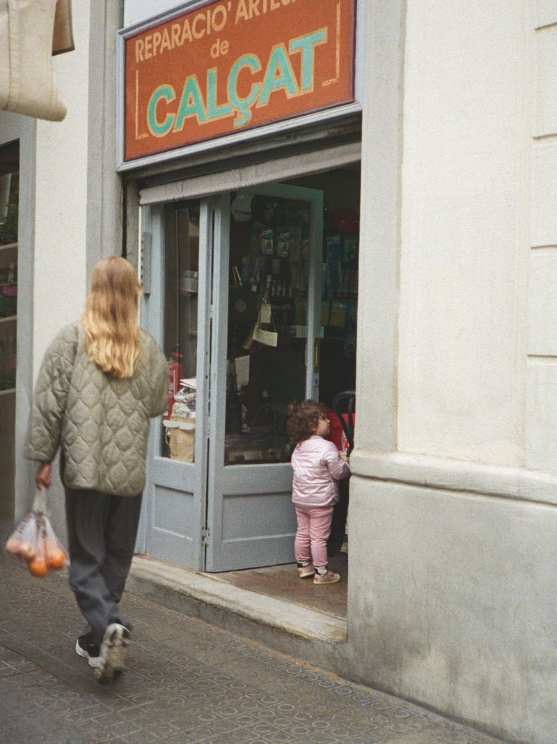 Woman walking away on a street carrying a bag of oranges past a storefront with a small child standing inside
