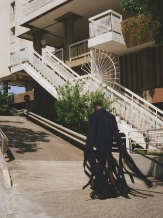 Model wearing a dark coat with long ribbon fringe walking near white outdoor stairs and apartment building