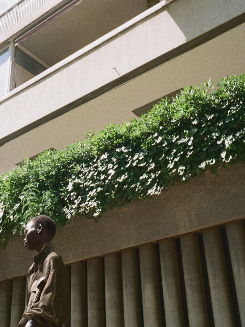 Model standing outdoors near a concrete building balcony filled with lush green foliage under bright sunlight.