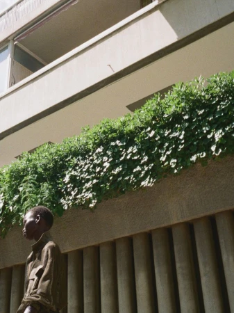 Model standing outdoors near a concrete building balcony filled with lush green foliage under bright sunlight.