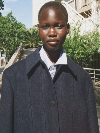 Model wearing a dark wool coat and light button-down shirt outdoors near leafy trees and architectural stairs.