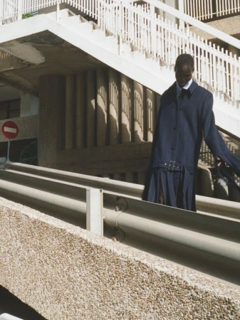 Model wearing a long dark blue coat standing outdoors near a concrete structure and white railings.