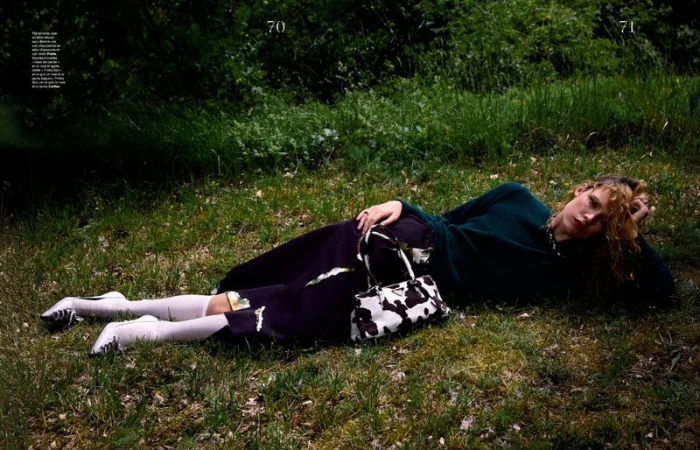Model lying on grass wearing a dark green top, dark skirt, white stockings, and holding a cow-print handbag.