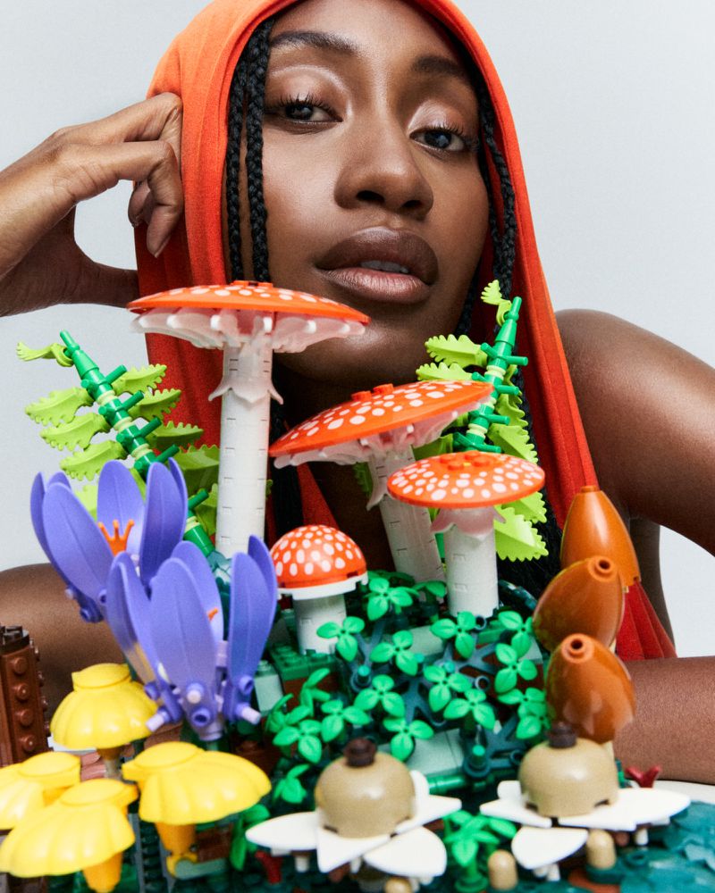 Model wearing orange hooded top behind a colorful arrangement of toy mushrooms and flowers