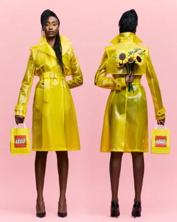 Model wearing a glossy yellow vinyl trench coat holding a Lego bag and sunflowers against a solid pink background.