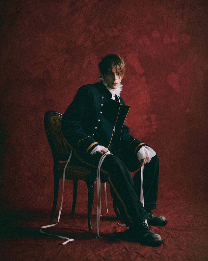 Corey Fogelmanis seated on an antique chair against a textured dark red backdrop wearing a dark military-style jacket.