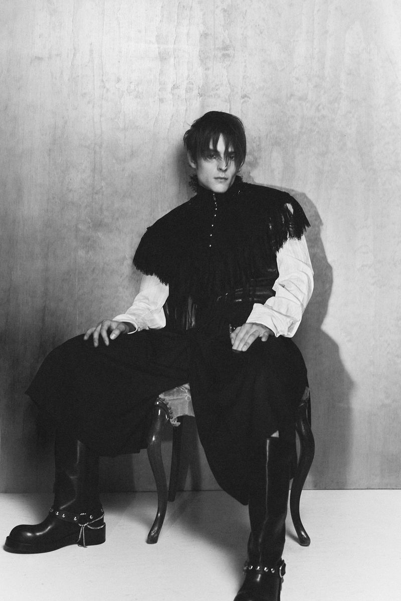 Monochrome portrait of a man seated on a chair wearing a dark collared top, white shirt, and tall boots against a textured background.