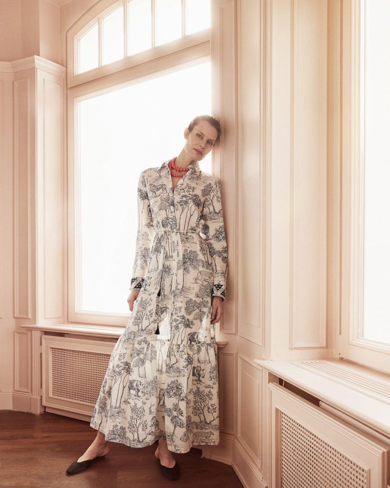 Fashion shot of woman in long-sleeve dress with toile pattern, posing in a corner of a light-peach coloured room, with large window