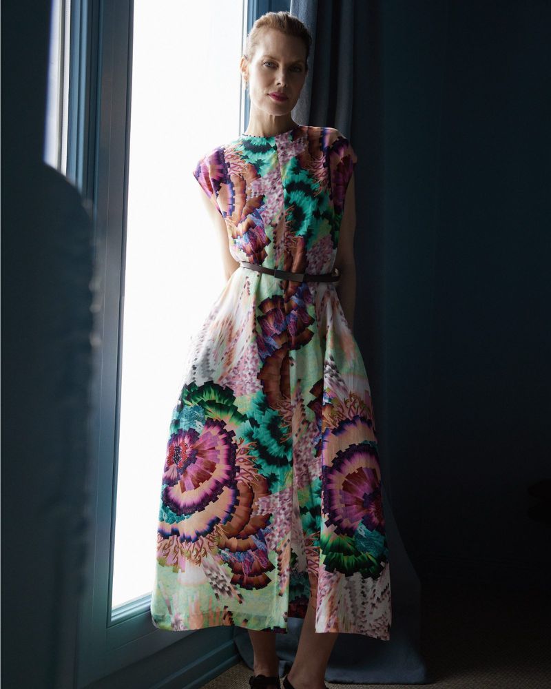 Indoor portrait of a blonde adult woman wearing a floral-patterned midi dress with a belt, in front of a window with dark curtains.