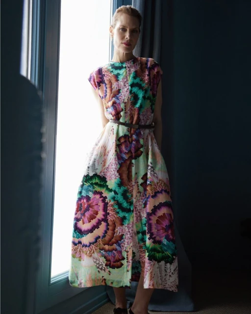 Indoor portrait of a blonde adult woman wearing a floral-patterned midi dress with a belt, in front of a window with dark curtains.