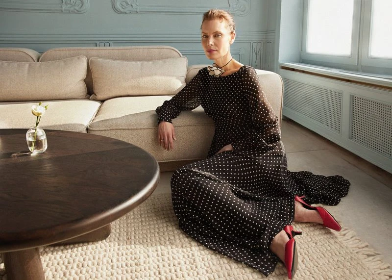Woman in polka-dot dress sits on rug in neutral toned room near couch and round table with flower. Red high heel shoes.