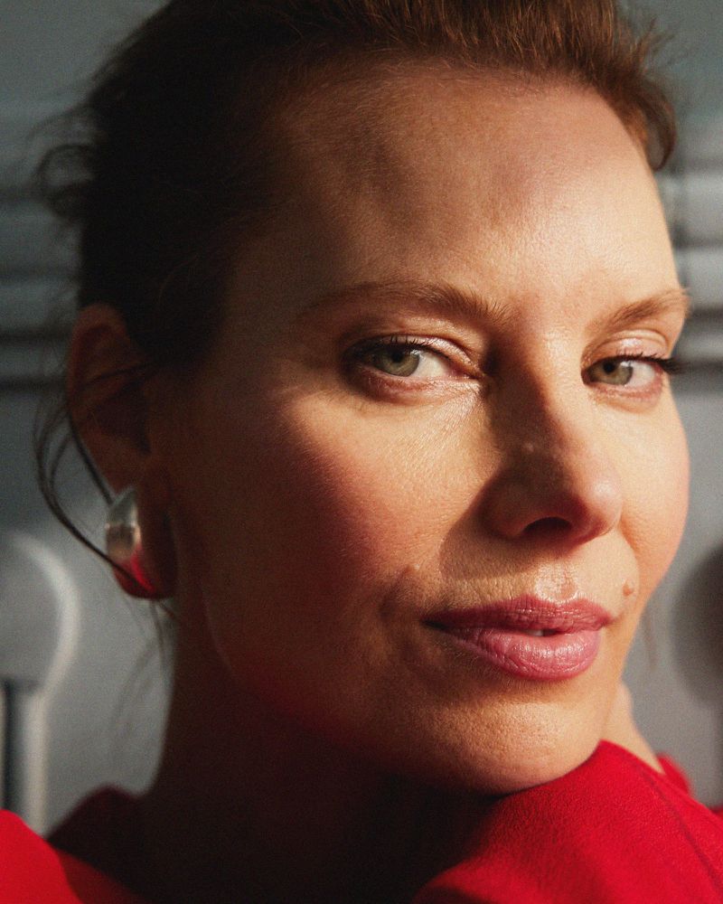 Close-up portrait of an adult female model with light skin, wearing a red blouse, with a silver earring, and gazing directly at the camera.