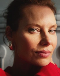 Close-up portrait of a woman wearing a red top with visible silver hoop earrings against a neutral grey wall.