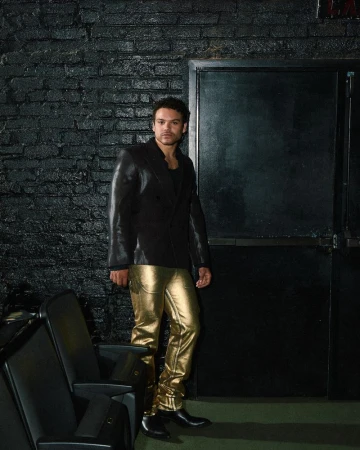 Josh Rivera wearing a black blazer and shiny gold pants standing in a dark interior space with theatre seats
