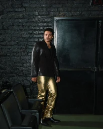 Josh Rivera wearing a black blazer and shiny gold pants standing in a dark interior space with theatre seats