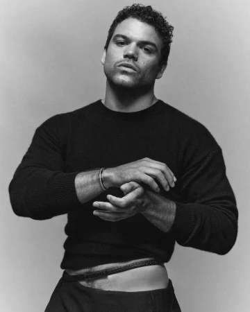 Portrait of Josh Rivera adjusting his bracelet, wearing a black sweater against a neutral background