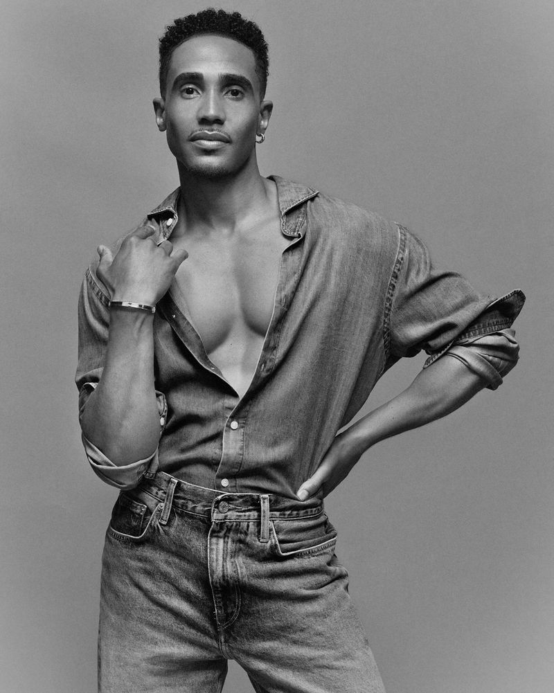 Black-and-white portrait of Jonathan Burke wearing an open denim shirt and jeans against a plain grey studio backdrop.