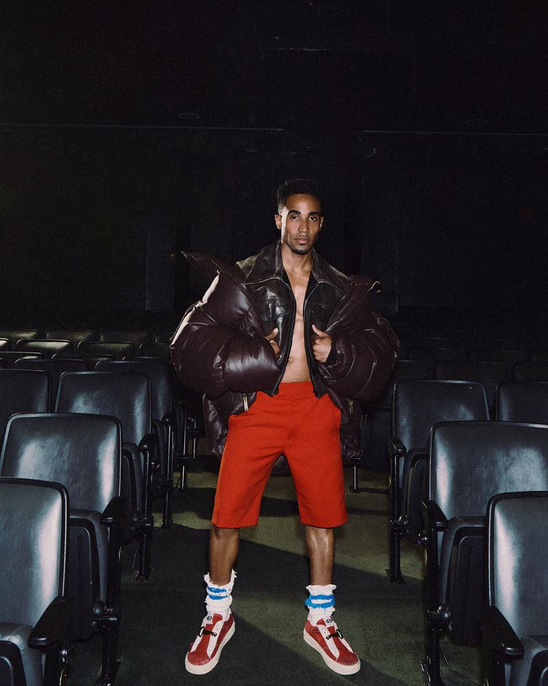 Man wearing a dark puffy jacket and red shorts standing in a dark movie theater with empty seats.