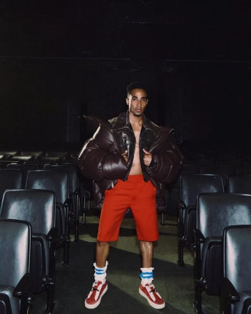 Man wearing a dark puffy jacket and red shorts standing in a dark movie theater with empty seats.