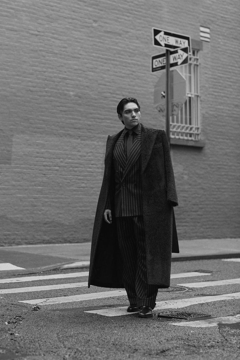 Man wearing a pinstripe suit and long dark coat walking on a city street near a one-way sign